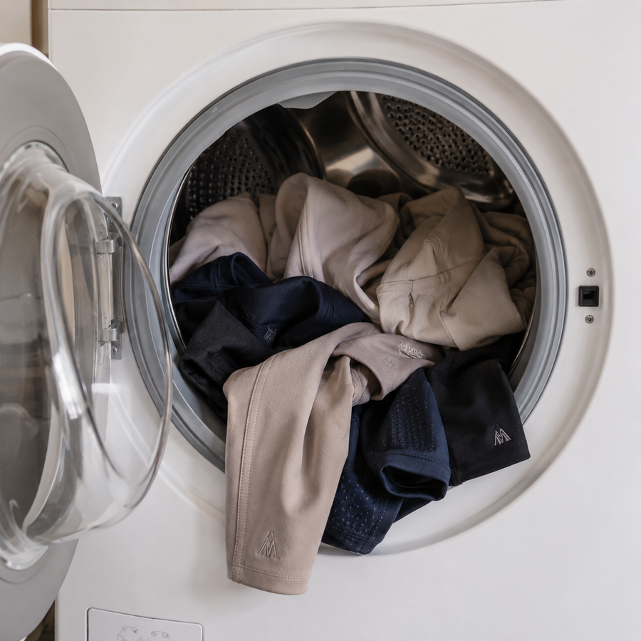 Riding breeches and equestrian base layers inside an open washing machine for a guide on how to wash riding clothes properly.